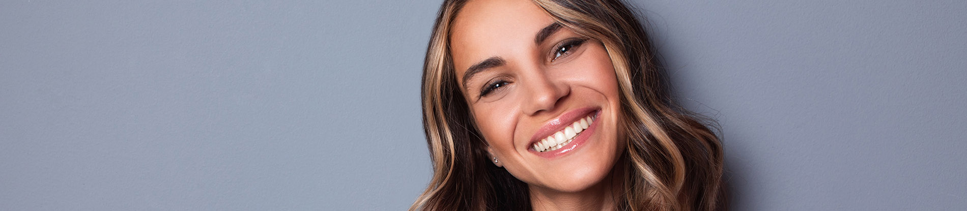 The image is a photograph of a woman with light skin, smiling at the camera. She appears to be in her late twenties or early thirties and has long hair. Her eyes are looking directly at the camera, and she is holding up her index finger near her mouth as if she s making a point or emphasizing something. The background is plain and light-colored, which suggests that this could be a stock photo used for various purposes such as advertising, personal branding, or lifestyle content.