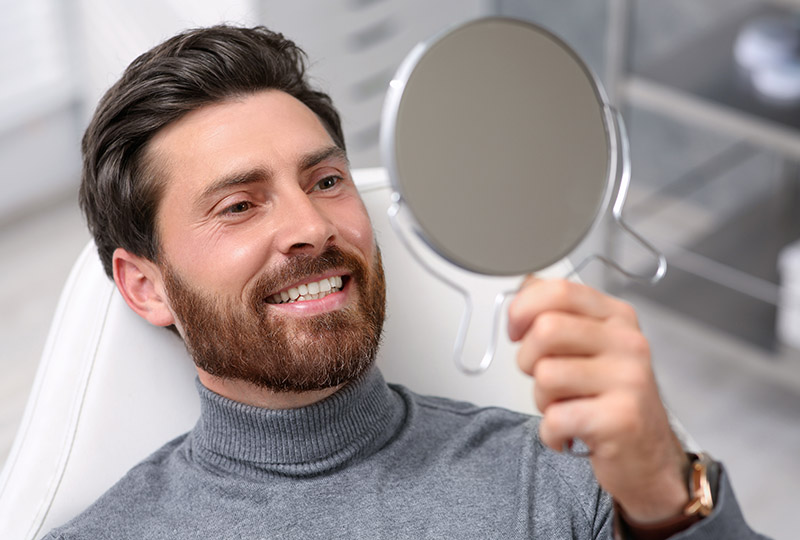 A man with facial hair smiling at his reflection in a magnifying mirror while holding the mirror.