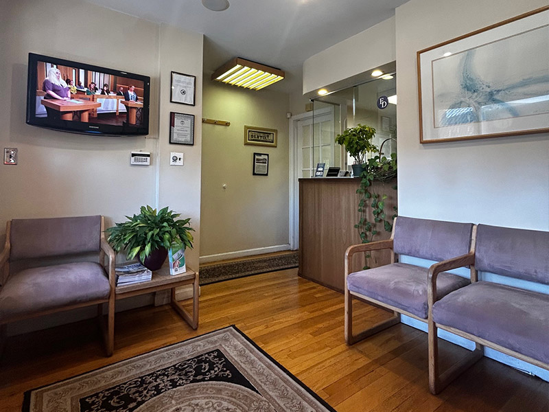 The image depicts an interior space with a waiting area featuring two chairs, a television monitor displaying content, a wall-mounted framed picture, and a potted plant on a floor stand.