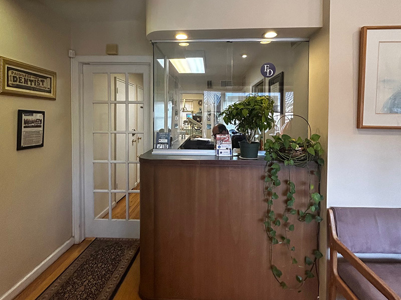 The image shows an interior view of a reception area with a wooden desk, a potted plant on the floor, and a framed picture on the wall.