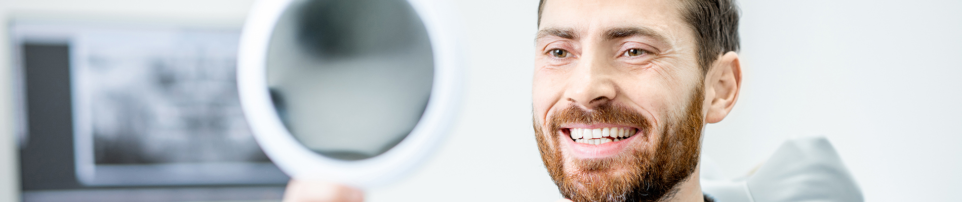 The image shows a man smiling at the camera, with his reflection visible in a mirror behind him. He appears to be indoors, possibly in an office setting, as suggested by the presence of a desk with papers on it and a computer monitor in the background.