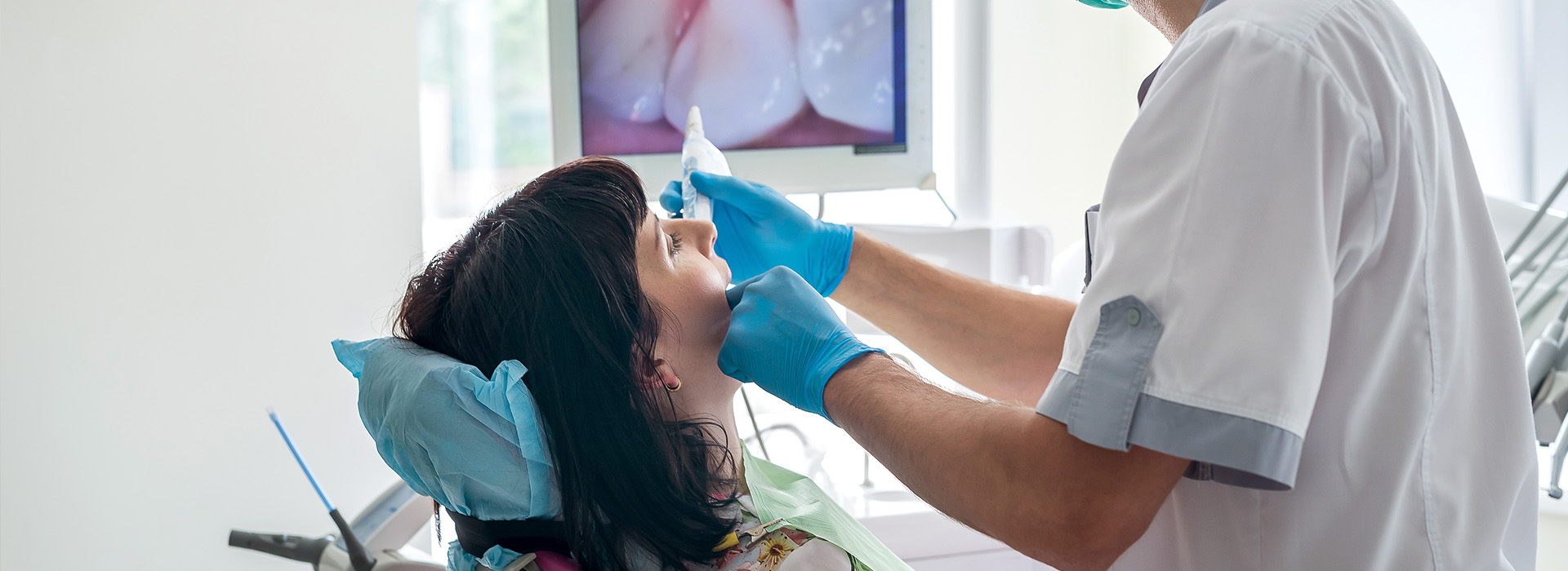 The image features a dental professional performing a procedure on a patient s mouth, with the professional using a dental instrument, while the patient appears to be seated in a dental chair and wearing a blue surgical mask.
