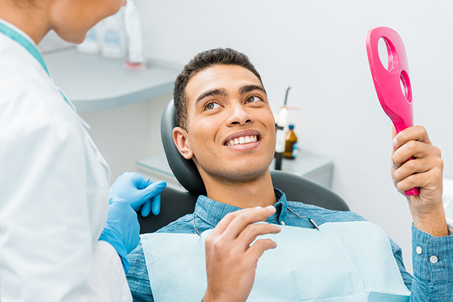 A dentist is sitting in a dental chair with a smile on his face while holding a pink object, possibly a toothbrush or dental tool, and being attended to by two dental professionals who appear to be assisting him during a dental appointment.