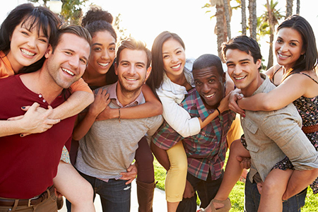 The image shows a group of people, likely friends or family, posing together for a photograph with smiles on their faces, standing outdoors during daylight with palm trees visible in the background.